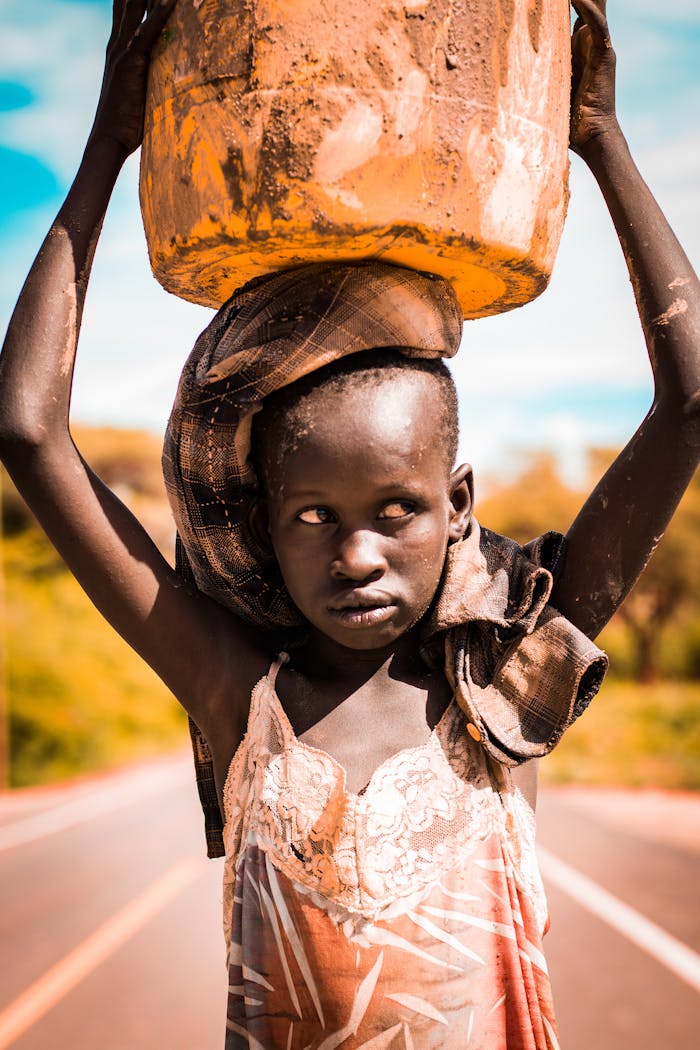 A young girl balances a water container on her head, walking on a road in rural Kenya.