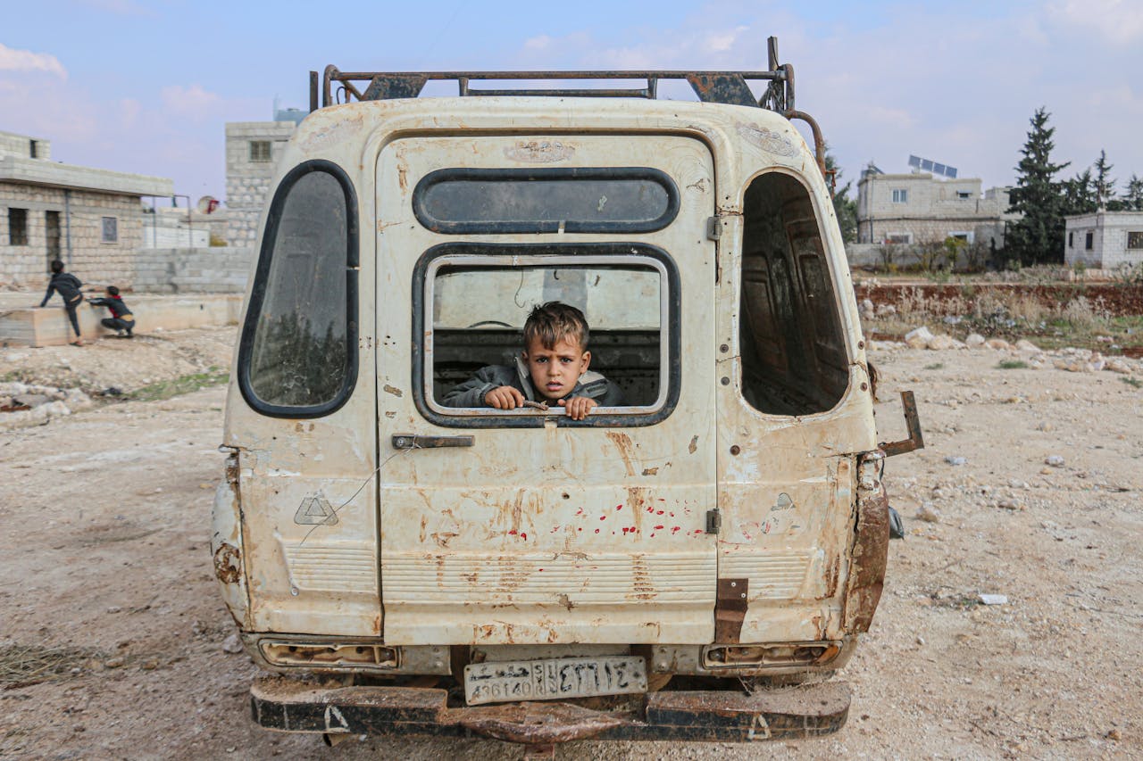 heros-img A young boy peers from an abandoned van in the desolate streets of Idlib, Syria, amidst war devastation.