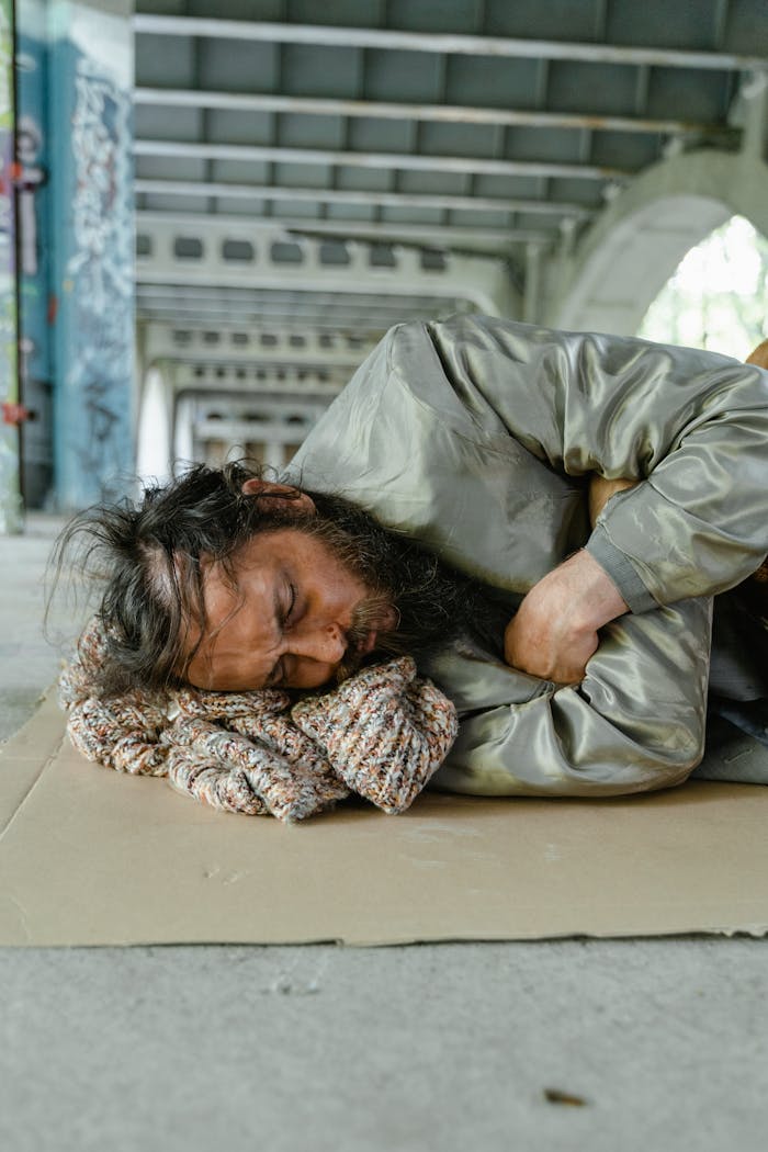 our-story A homeless man with a beard is sleeping on a cardboard under a bridge, highlighting urban poverty.